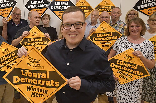 Jack Davies holding a Lib Dem diamond poster saying "winning here" with a group of members behind